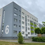 Frisch sanierte Fassade eines mehrgeschossigen Wohnhauses mit großflächiger Hausnummer „6“ in Weiß auf hellgrauem Hintergrund sowie akzentuierten grünen Fassadenelementen. Verweis: Fassadeninstandsetzung Magdalenenluster Weg, Güstrow