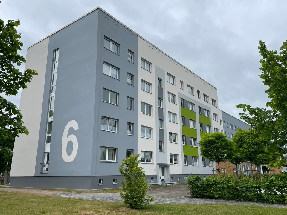 Frisch sanierte Fassade eines mehrgeschossigen Wohnhauses mit großflächiger Hausnummer „6“ in Weiß auf hellgrauem Hintergrund sowie akzentuierten grünen Fassadenelementen. Verweis: Fassadeninstandsetzung Magdalenenluster Weg, Güstrow