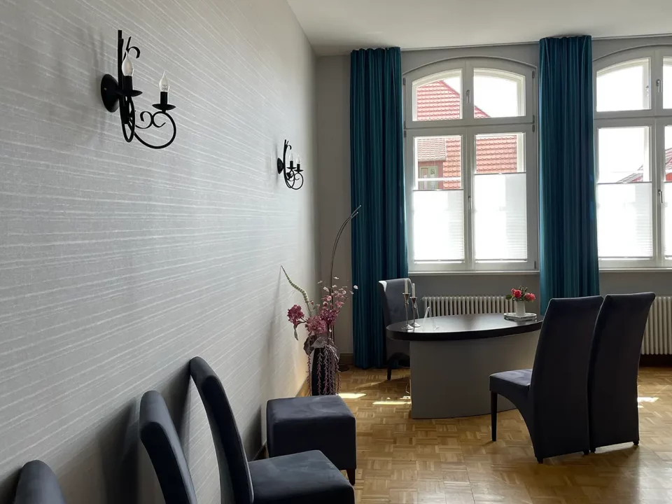 Blick auf das helle, modern renovierte Trauzimmer mit großen Rundbogenfenstern, dunkelblauen Vorhängen und einem stilvollen Schreibtisch vor einem Heizkörper.