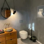 Modern gestaltetes Gäste-WC mit gespachtelten, fugenlosen Wänden in Betonoptik, hängendem WC und individuell gefliestem Waschbecken auf Holzkommode.