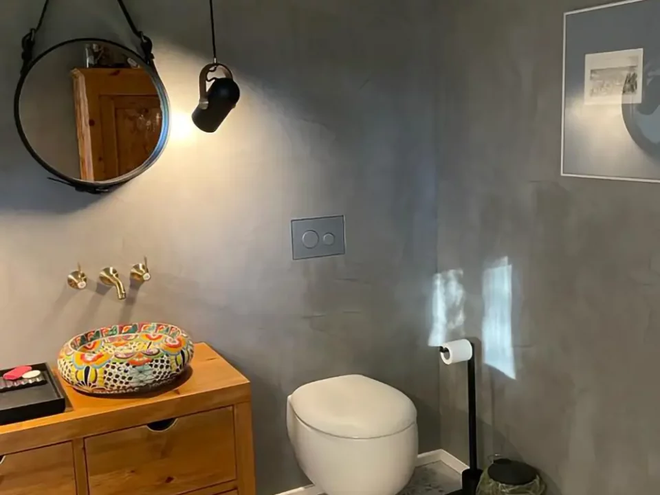Modern gestaltetes Gäste-WC mit gespachtelten, fugenlosen Wänden in Betonoptik, hängendem WC und individuell gefliestem Waschbecken auf Holzkommode.
