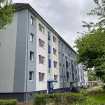 Perspektive entlang des sanierten Gebäudes – zurückhaltende Farbwahl in Grau und Weiß mit vertikalen Farbflächen, harmonisch eingebettet ins grüne Wohnumfeld.